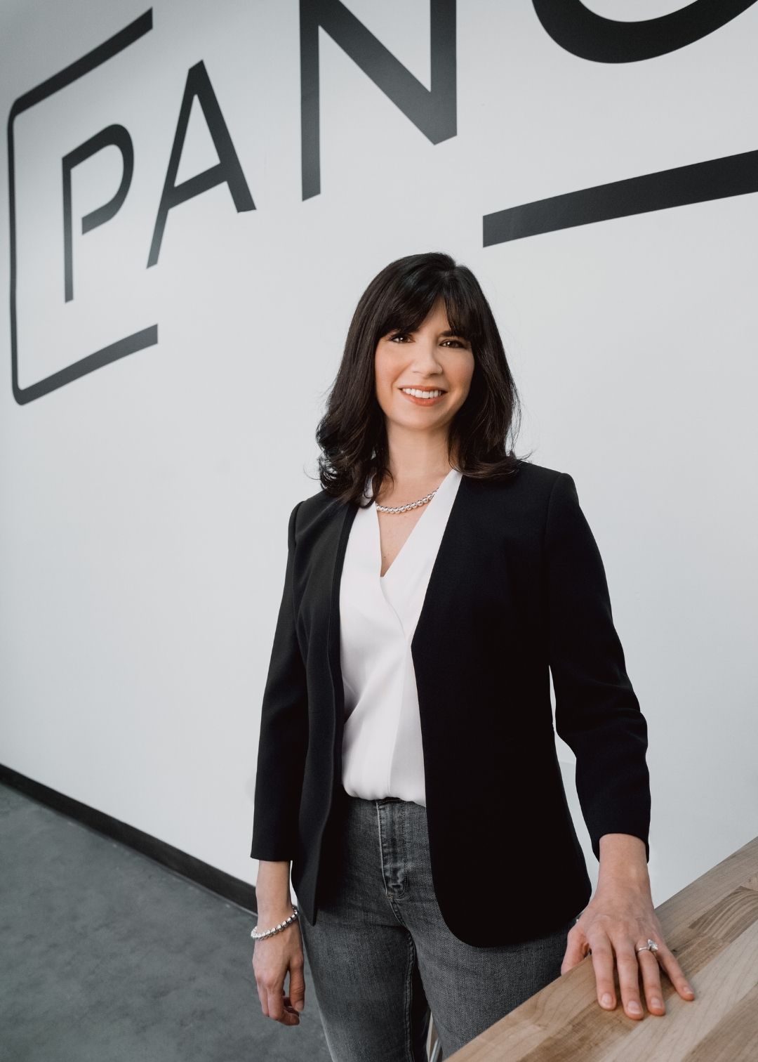 A woman in a black blazer and white blouse stands indoors, smiling, with part of a large wall sign reading "PAN" in the background—perfect for testimonials or professional profiles.